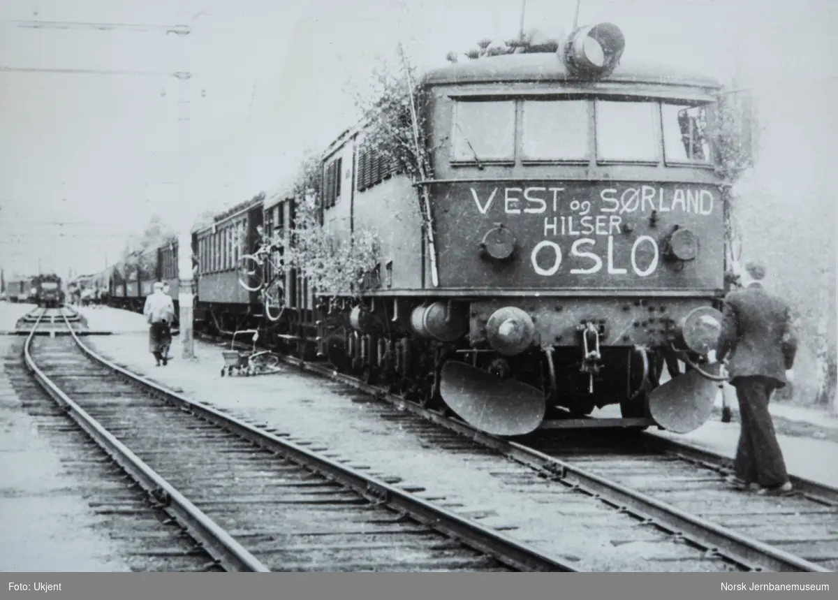 Elektrisk lokomotiv i tog 706 pyntet løv og påskrevet "Vest og Sørland hilser Oslo" på Hjuksebø stasjon.