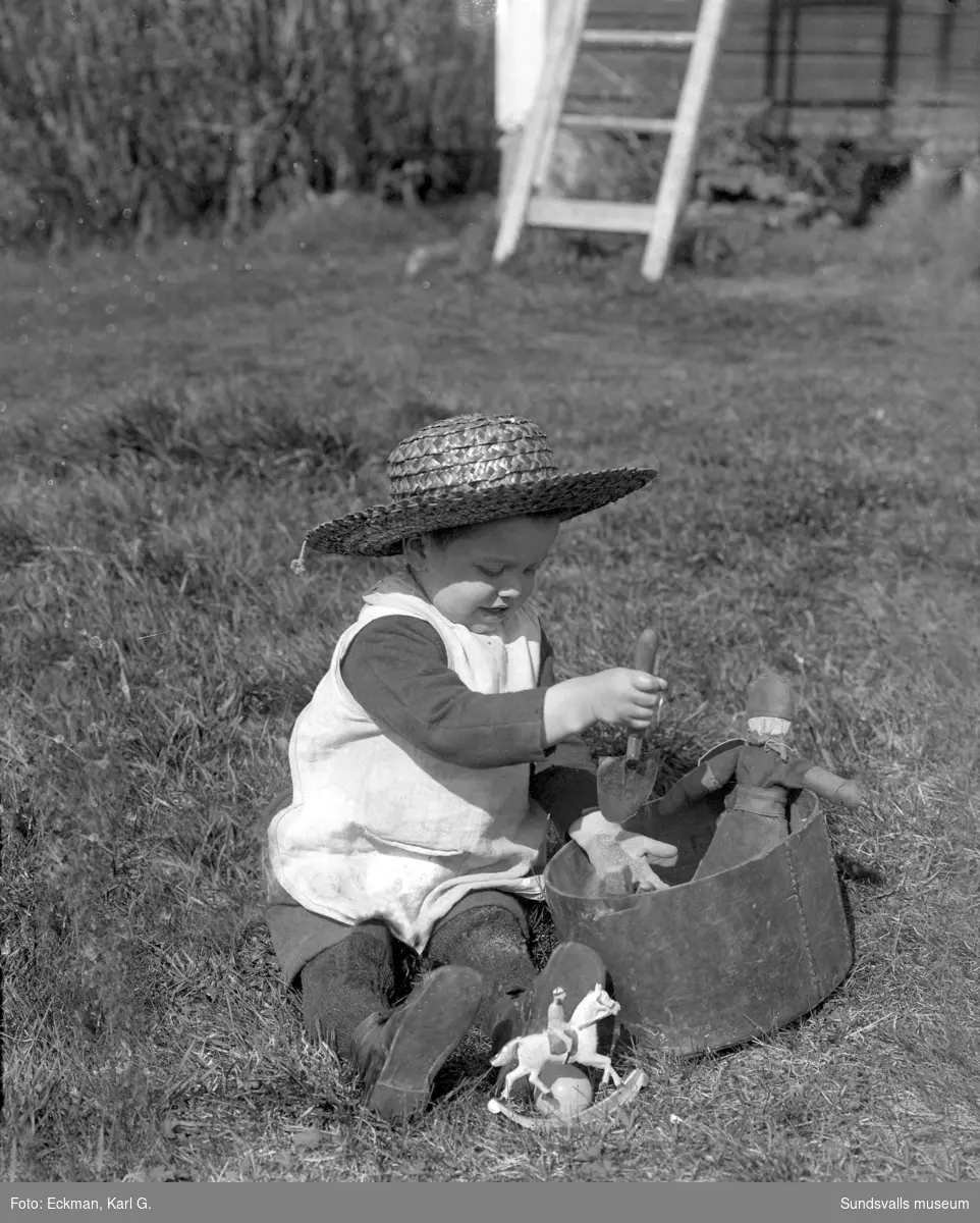Ett barn, möjligen Rakel eller Ulla Eckman, med halmhatt leker med sand och en spade med docka och leksakshäst intill. Ur fotograf Karl Eckmans samling.