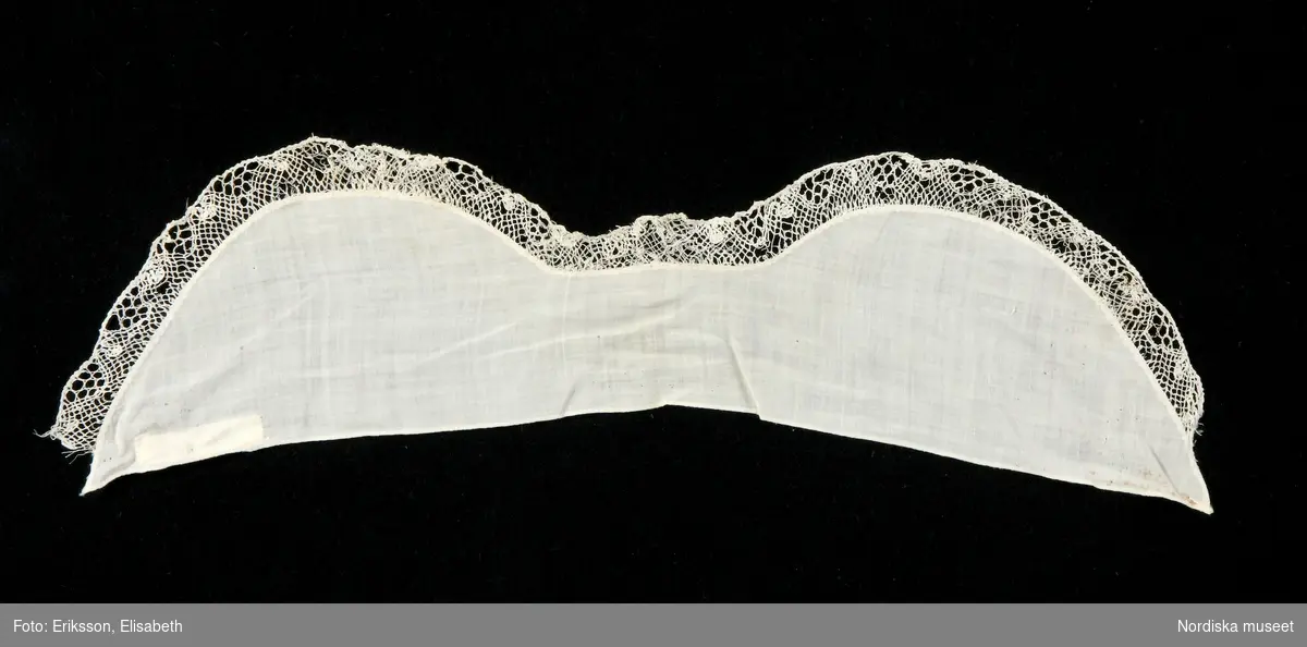 Stycke av tunn vit bomullslärft, formad i 2 bågar, handfållad och med fastsydd gles knypplad spets i kanten, 2 cm bred.
/Berit Eldvik 2010-02-19