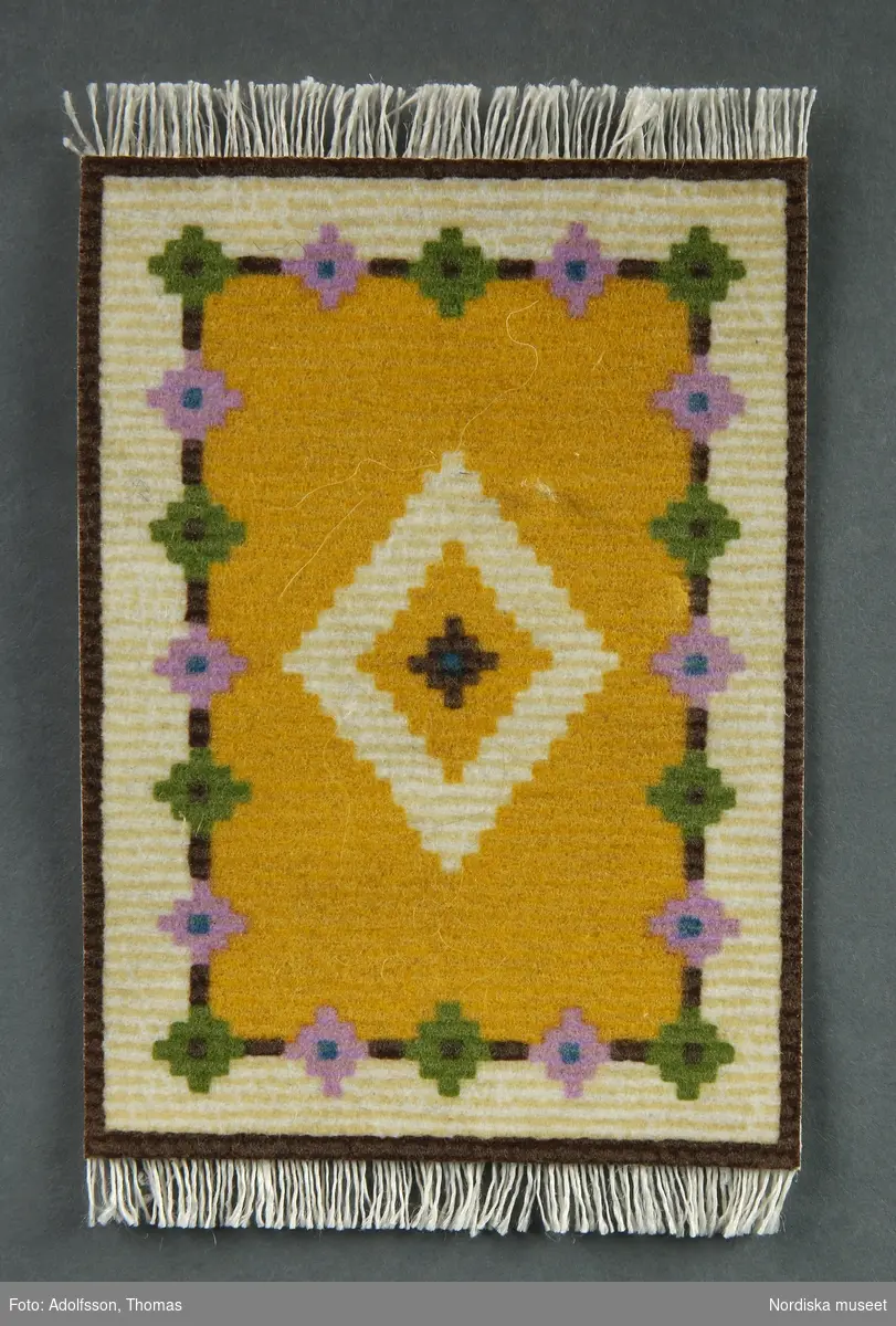 Två matsalsmattor i miniatyr, den ena a) föreställer en röllakansmatta och den andra b) en orientalisk matta. Röllakansmattan går i färgerna gult, vitt, grönt och rosa och ska ligga mitt i rummet, under matgruppen (NM.0333310 a-e). Den stämmer färgmässigt väl överens med det gulvita möblemanget. Den orientaliska mattan är röd, brun och grön och ligger under sittgruppen (NM.0333310 f-h) vid det lilla pelarbordet nära fönstret vid terrassen.