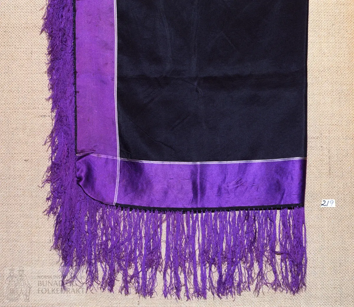 Svart silketørkle med fiolette stripar langs kantane. Påsette fiolette silkefrynser, 13 cm lange.  Storleik:  78 x 81 cm.