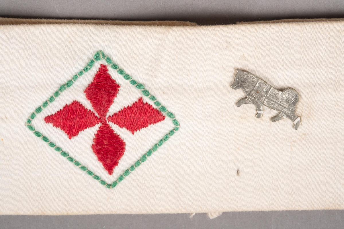 Et armbind i hvit bomull med rødt kors brodert i midten. Ved korset er det påsydd et merke av en hund.
