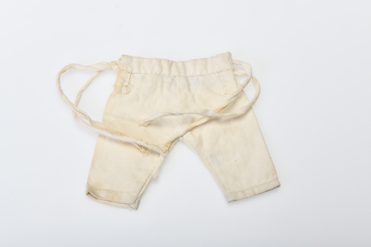 En bukse/underbukse til dukke sydd av hvitt bomullsstoff. Den har knelange bein og har knytting i siden (to bånd i hver side).