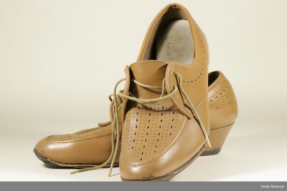 Brune damesko i skin. Skoen har en liten hel. Det er skolisser i brunt tøy, for å knytte skoen. Skoen er dekorert med små hull på midten av skoen, og langs kanten. Skoene er delvis slitt inni og på sålene. Skoen har skostørrelse 39. Skoene er av finsk fabrikat. 