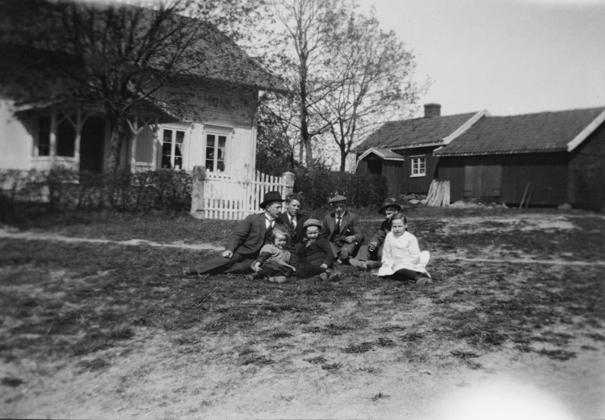 Gruppebilde med sju voksne og barn i gresset foran to gårdsbygninger