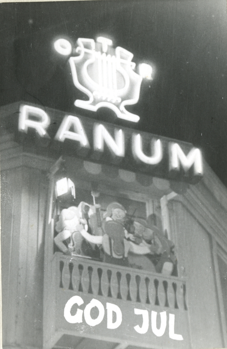 Takpynten på butikken til Ranum a/s i Olav Trygvassons gate 18. Vi ser butikkens logo i neonlys på toppen av taket. Under "Ranum" i neonbokstaver. På den lillebalkongen er det satt opp flere nisser som spiller instrumenter og hengt opp en lampe. Under balkongen, kun på fotografeit, er det skrevet "God Jul".