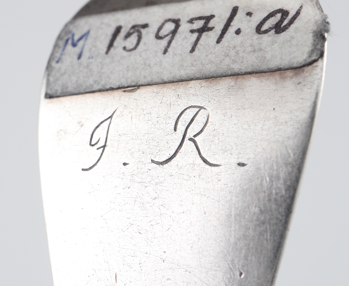 Gaffel av silver med uppåtböjd bak. 
Märkt med initialerna "J.R." på baksidan. Gaffeln har tre uddar, brett flat skaft med markerad ås på ovansidan. 
Gaffeln är helt slät och odekorerad. Stämplar på skaftets baksida.

Inskrivet i huvudbok 1955.