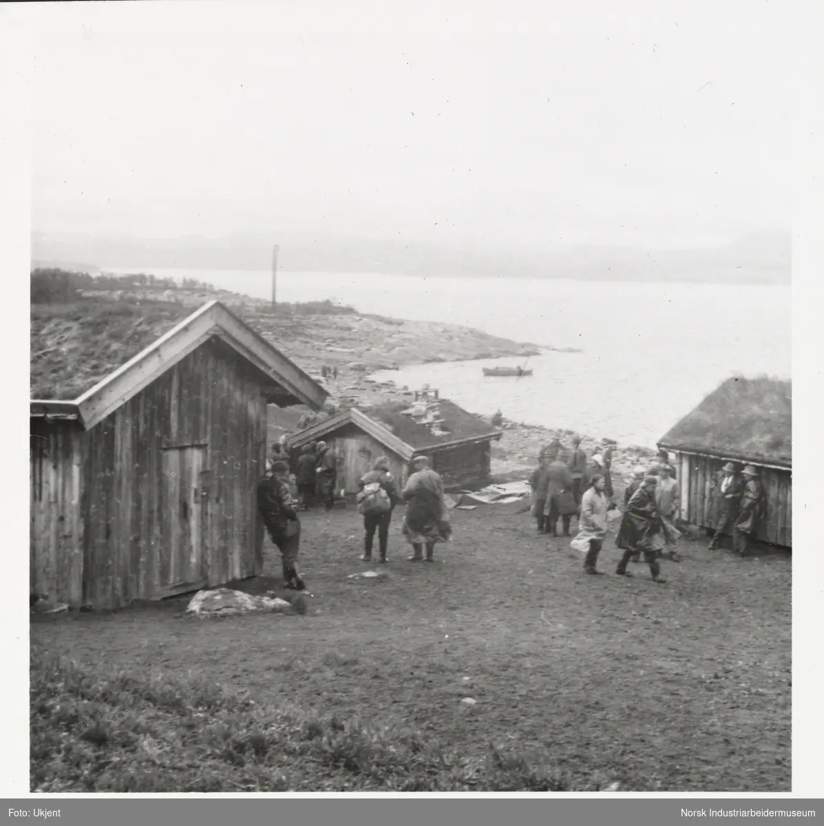 Mennesker står ved bygninger ved vannkanten til innsjøen Møsvatn. På vannet sees en snekke