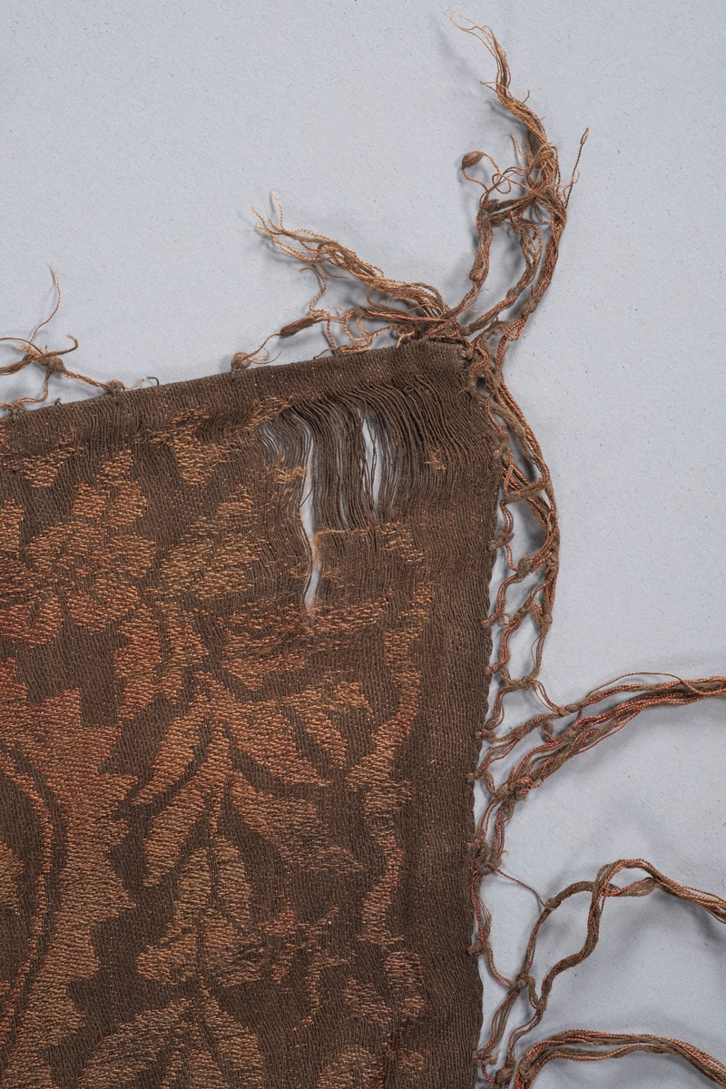 Rektangulært kobberfarget innslag sjal i bomull. På begge kortsidene er det en flettet kant med knyttede frynser ytterst. Sjalets underside er lysere enn oversiden.