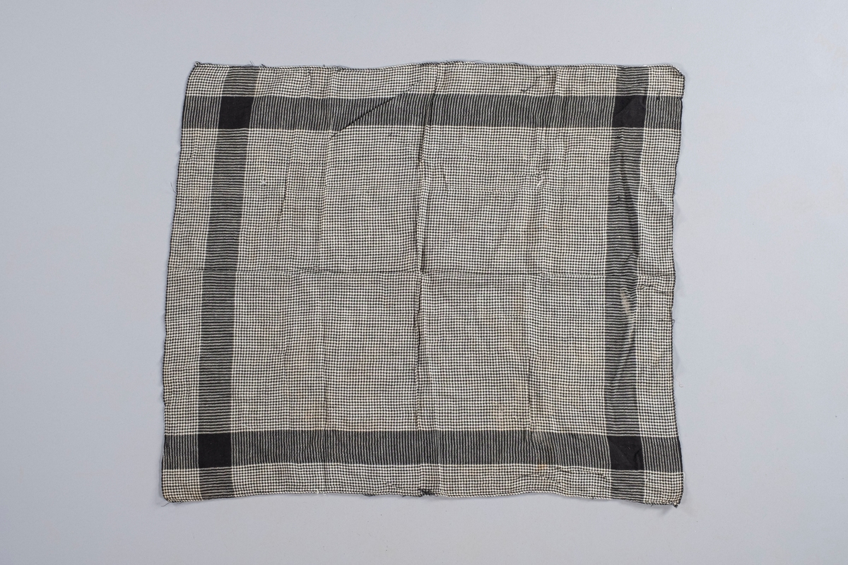 Rektangulært tørkle i svart-hvit bomullstekstil. Sydd for hånd i ene kanten. Motiver: ruter, striper og kvadrater.