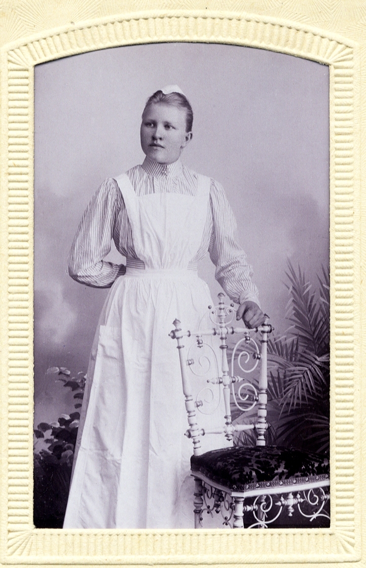 Foto av en kvinna i sjuksköterskeuniform (?) med hätta.
Helfigur, halvprofil. Ateljéfoto.