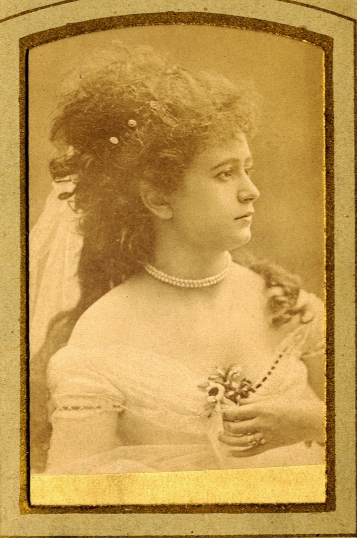 En kvinna klädd i urringad festklänning och halsband m.m.
Bröstbild, profil. Ateljéfoto.