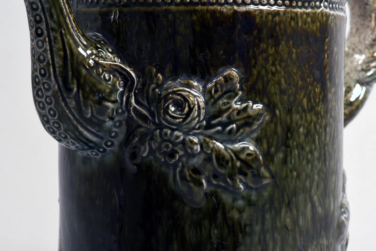 Kaffekanna av fajans med mörkgrön glasyr, försedd med pip och handtag. Kannan är cylindrisk och avsmalnande mot mynningen. Dekorerad med tre pärlstavsbårder runt kannan samt en bård längs handtaget. Vid den svängda dekorerade pipen är kannan dekorerad i relief med blad och rosor, likaså vid handtaget som är utformat som en gren.