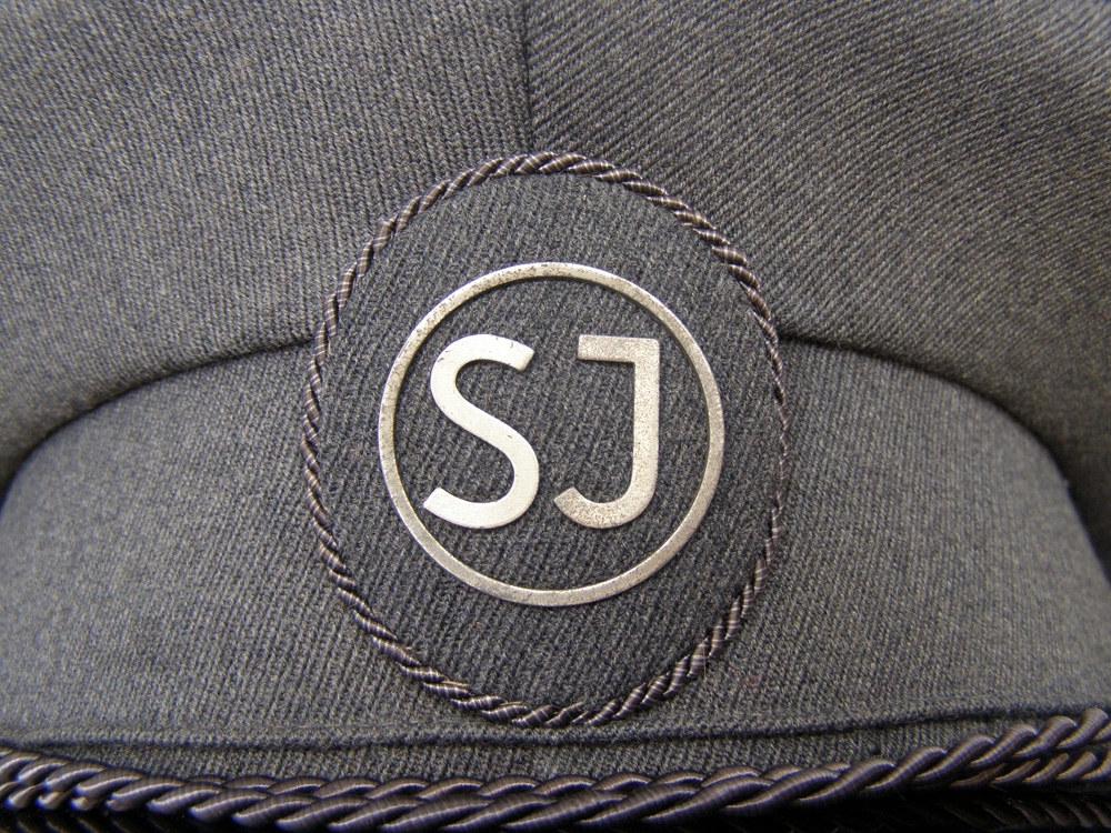 Skärmmössa av grått ylle, s k diagonaltyg, med grå stormträns och ovalt mössmärke klätt med grått tyg och bokstäverna SJ inom ring i silverfärgat metall.
Även grå mösstränsknapp med SJ:s logga.
