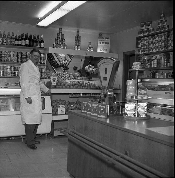 Köpman Åke Rahm i vit rock står i sin butik vid hyllor med konserver och disk med frukt.