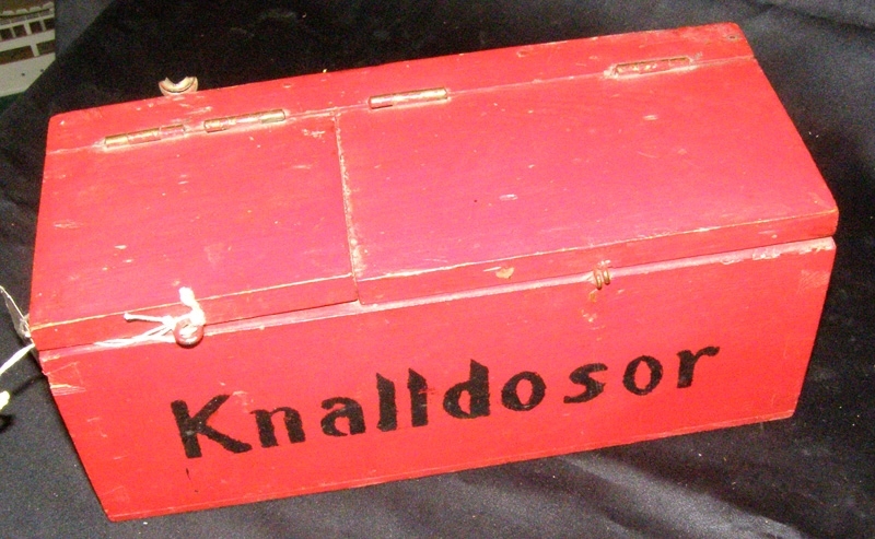 Rödmålad trälåda för knalldosor, med texten "Knalldosor" målat i svart på framsidan. Locket är tudelat. Lådan är tom.