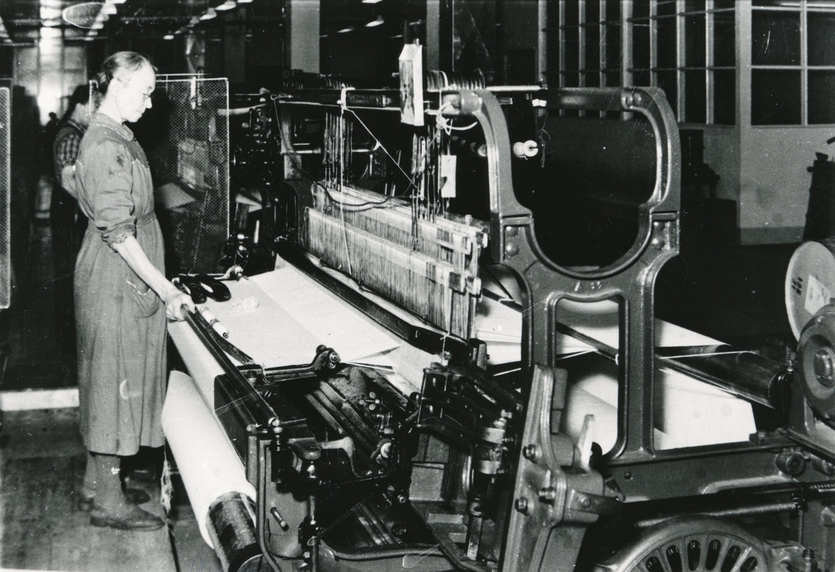 En kvinne står å jobber med en maskin som vever materiale i Devold fabrikken.