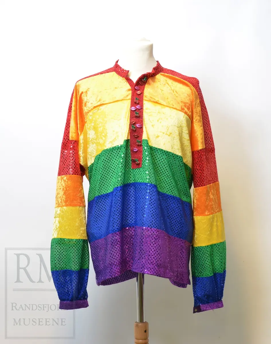 Skjorte i busserull-fasong sydd i regnbuefarget paljettstoff og fløyel. Dekorert med kirsebær- og leppe knapper, samt spensel i ryggen.