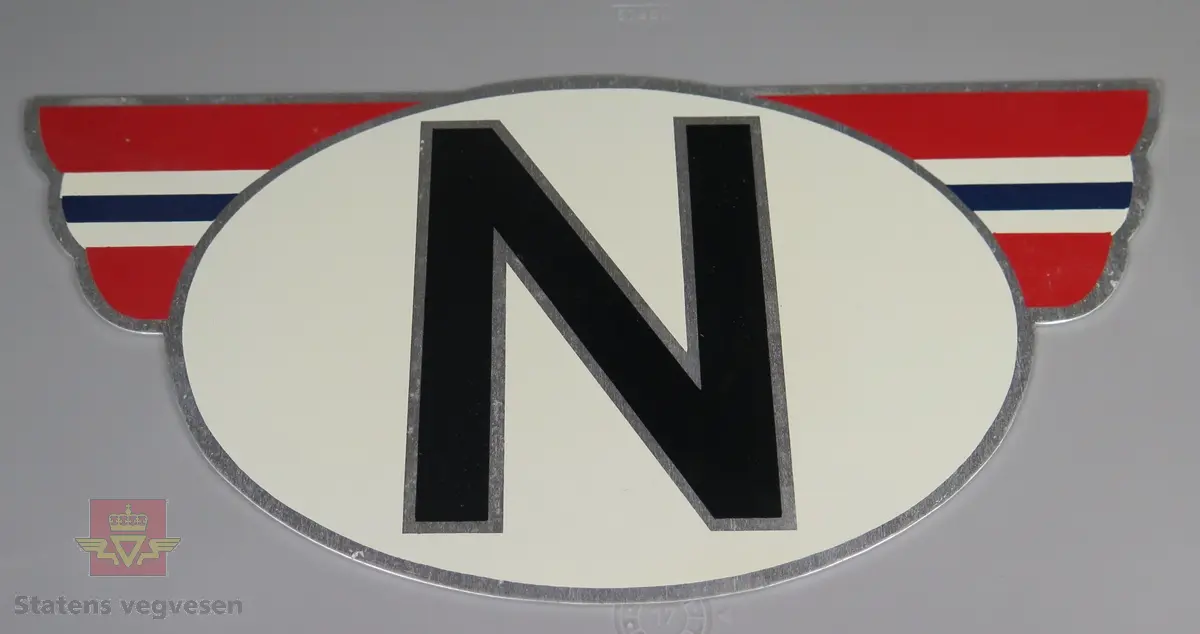 Skilt av aluminium. Bokstaven N i svart på hvit bunn omkranset av vinger i rødt, hvitt og blått. Emballasje med informasjon fra produsenten.