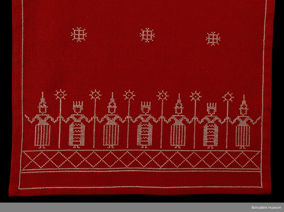 Vita korsstygn på röd botten, aidaväv. Stiliserade ljusbärare och snöstjärnor som motiv.