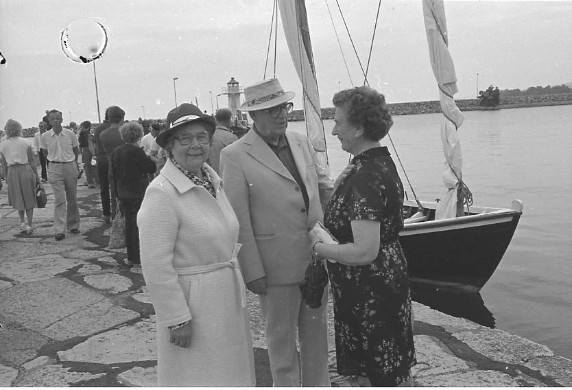 Maj och Tage Grennfelt står vid kajen i Gränna hamn och pratar med Ester Nordenankar. En snipa e motsv med segel ligger vid kaj och mycket folk i rörelse. Det är sommar.