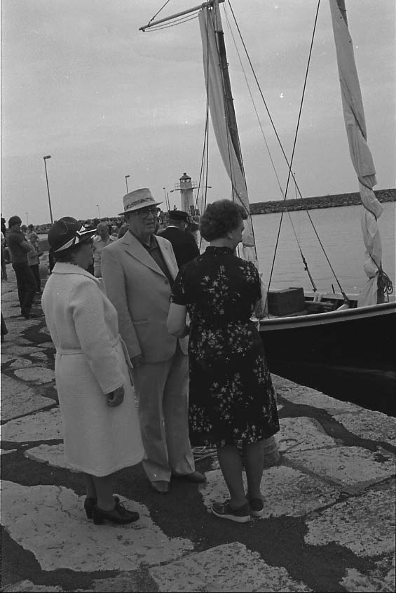 Maj och Tage Grennfelt står vid kajen i Gränna hamn och pratar med Ester Nordenankar. En snipa e motsv med segel ligger vid kaj och mycket folk i rörelse. Det är sommar.