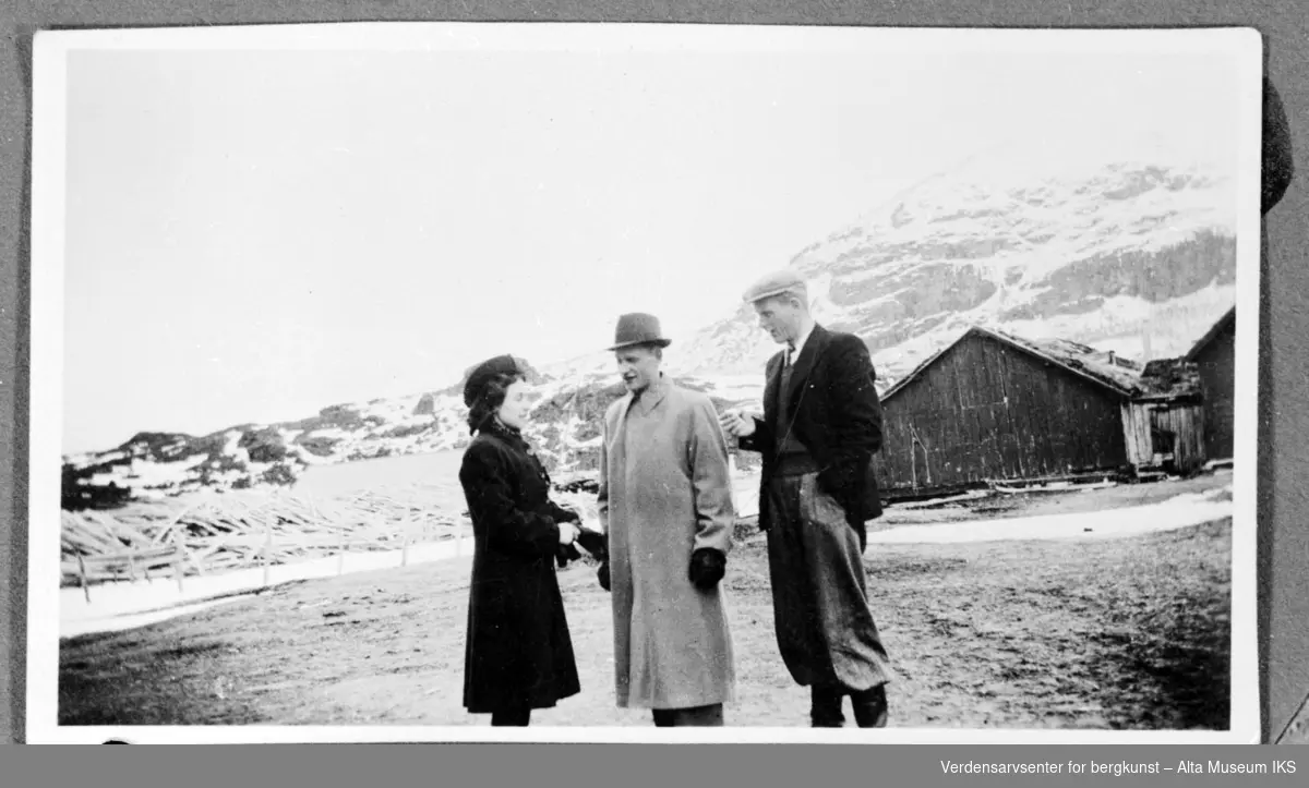 3 personer avbildet foran et tysk materiallager på kviby med et snødekket fjell i bakgrunn.