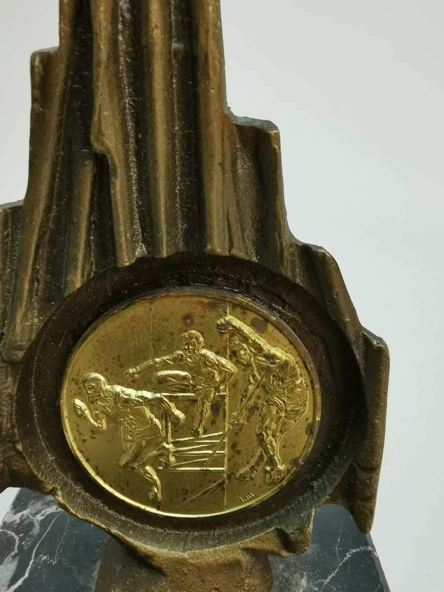 Medalje montert på bronse plate med mønster. Platen har en sokkel av svart marmor. Sokkelen er skadet  og mangler en bit i underkant.