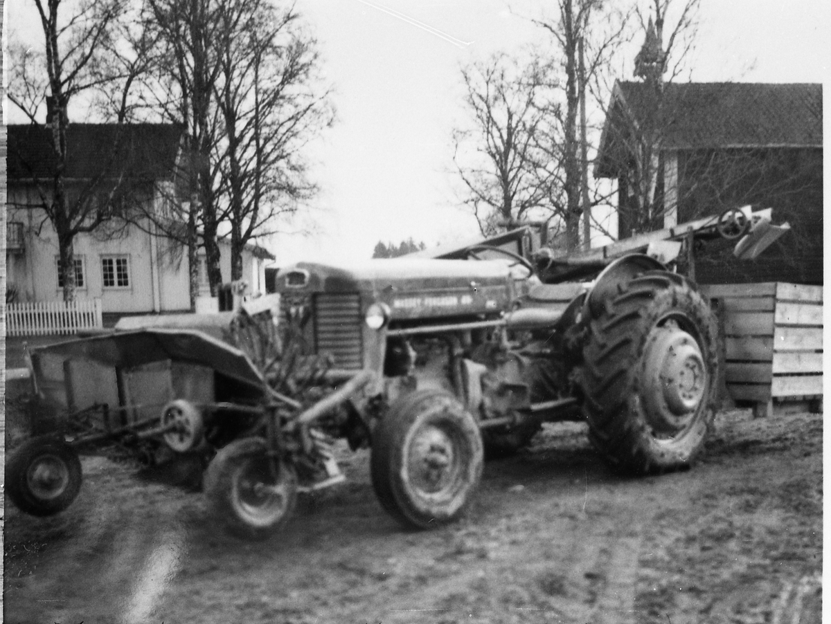 Avfotografert bilde av en traktor med potetopptaker som synes å være delvis frontmontert, men med transportbånd til potetkasse bak. Traktoren står på et gårdstun, og i bakgrunnen er et våningshus og et stabbur. Stedet er ikke identifisert.