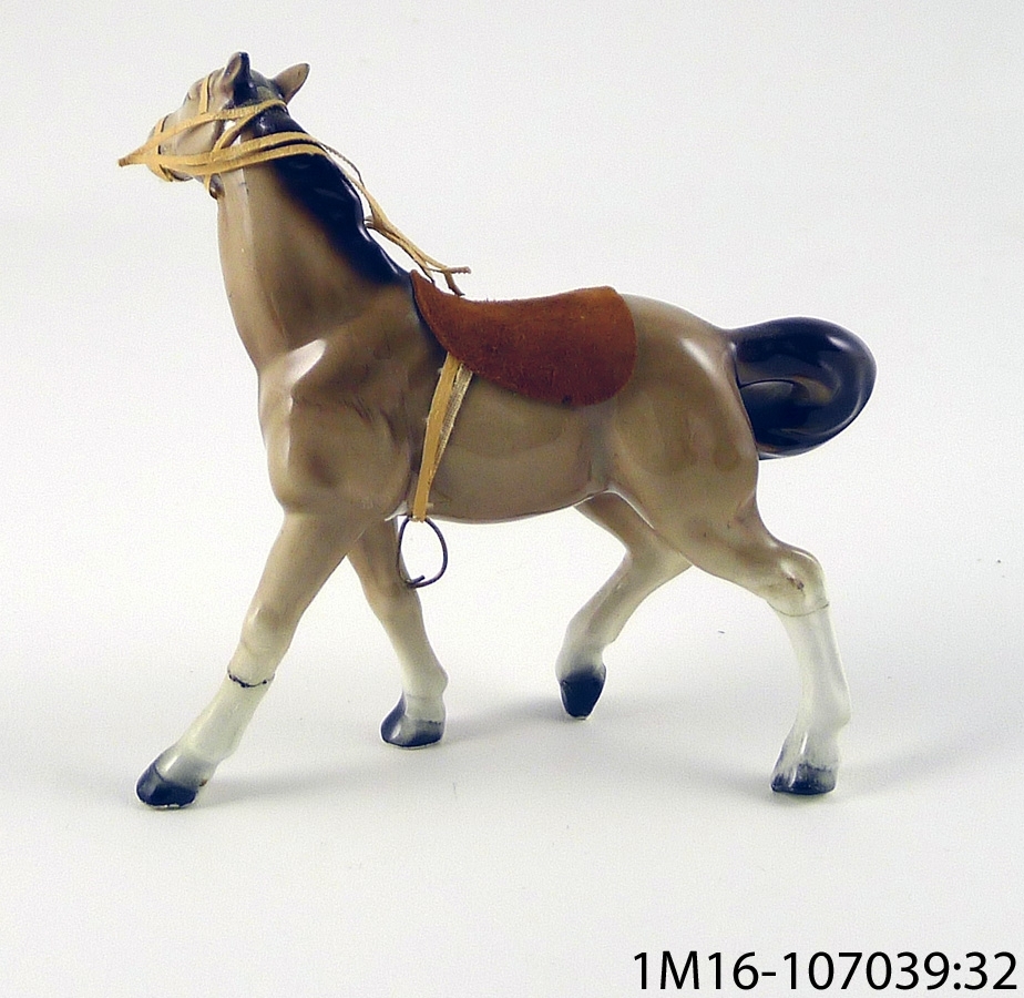 Häst i keramik, brun med sadel stigbyglar och remtyg. Hästen trasig, lagad med klister. Ena stigbygeln har lossnat.
