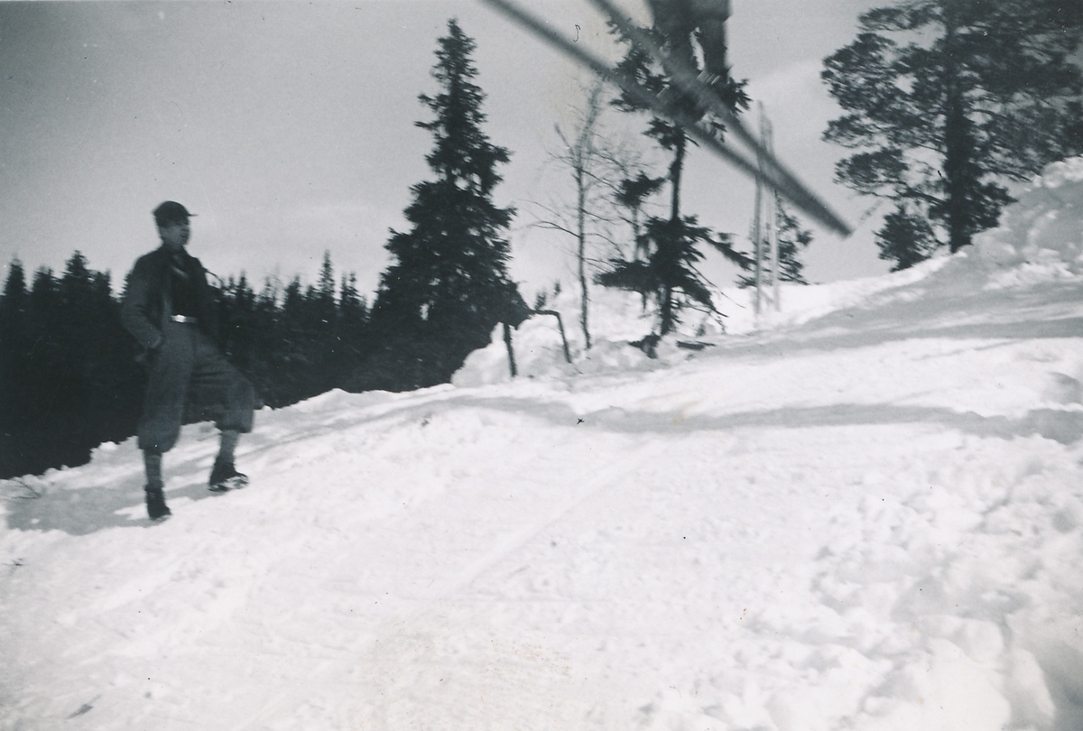 Hopping på ski. En person er midt i svevet, mens en mann står og ser på i ført eplenikkers og skyggelue.
