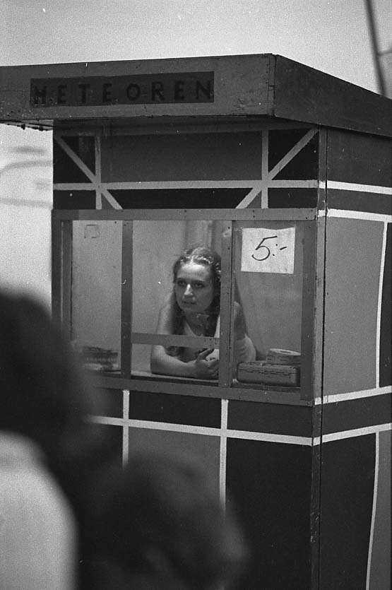En ung kvinna sitter i ett biljettstånd, förmodligen för en karusell e dyl som kostar "5:-". På dess tak syns texten: "Meteoren".