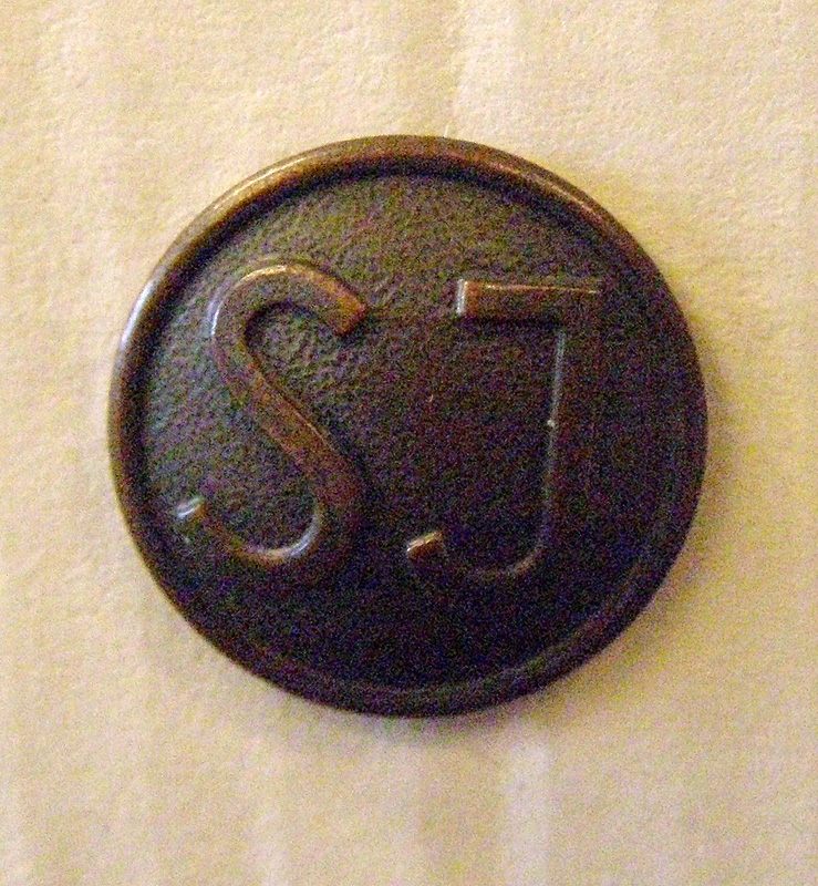 Brun mösstränsknapp av koppar med SJ:s logotyp i form av enkla initialer.