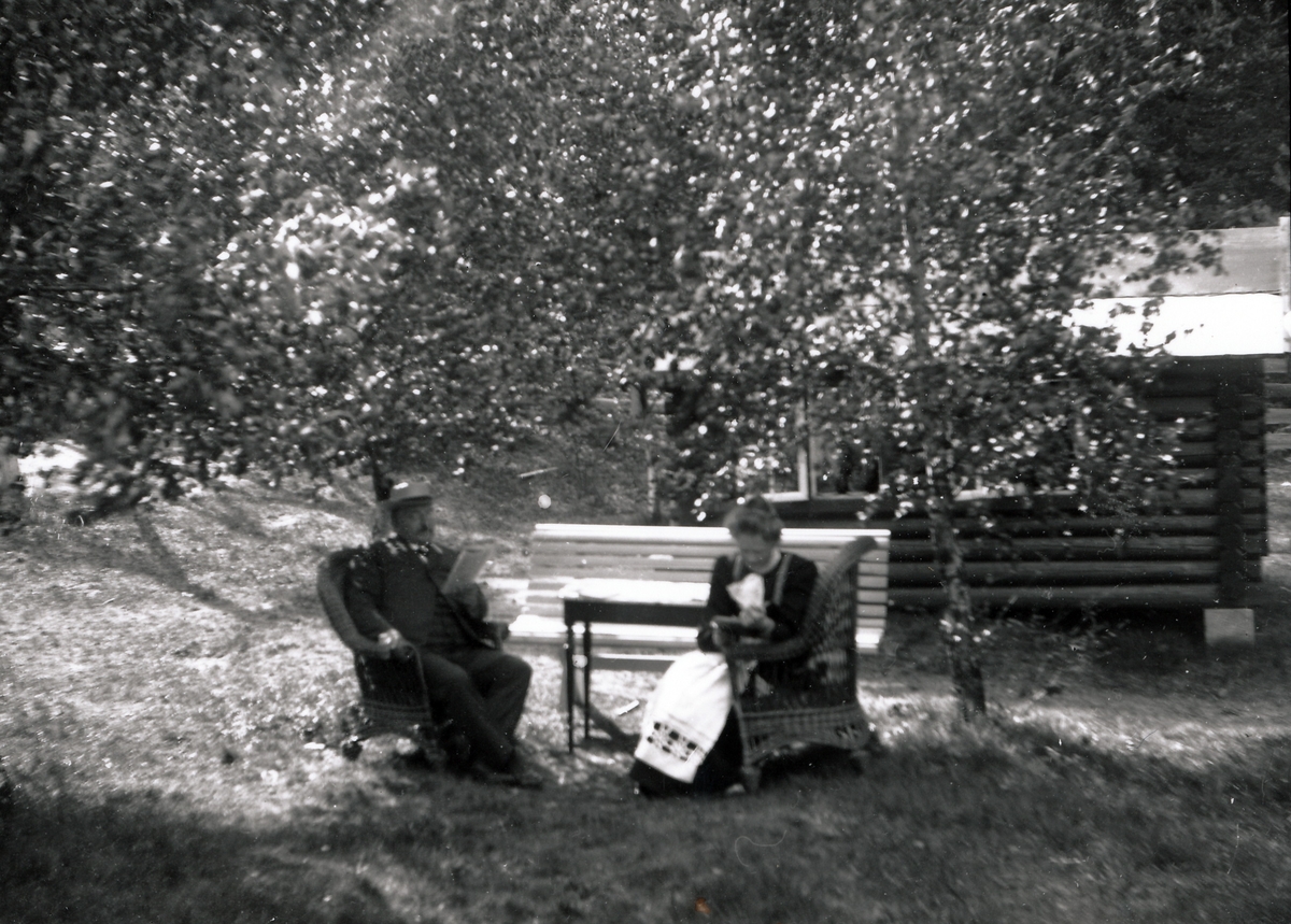 Bestyrer Wilhelm Rydgren og fru Nicoline Rydgren sitter i en hage. De sitter i kurvstoler ved et hagebord under bjørketrær.