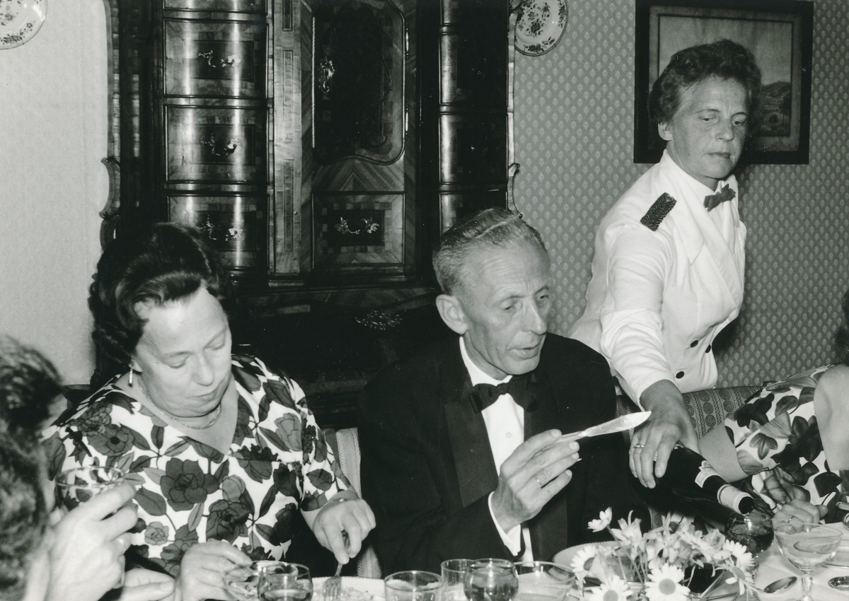 Kvinne og mann i festantrekk ved festbord, mens serveringsdame skjenker i vinglass.