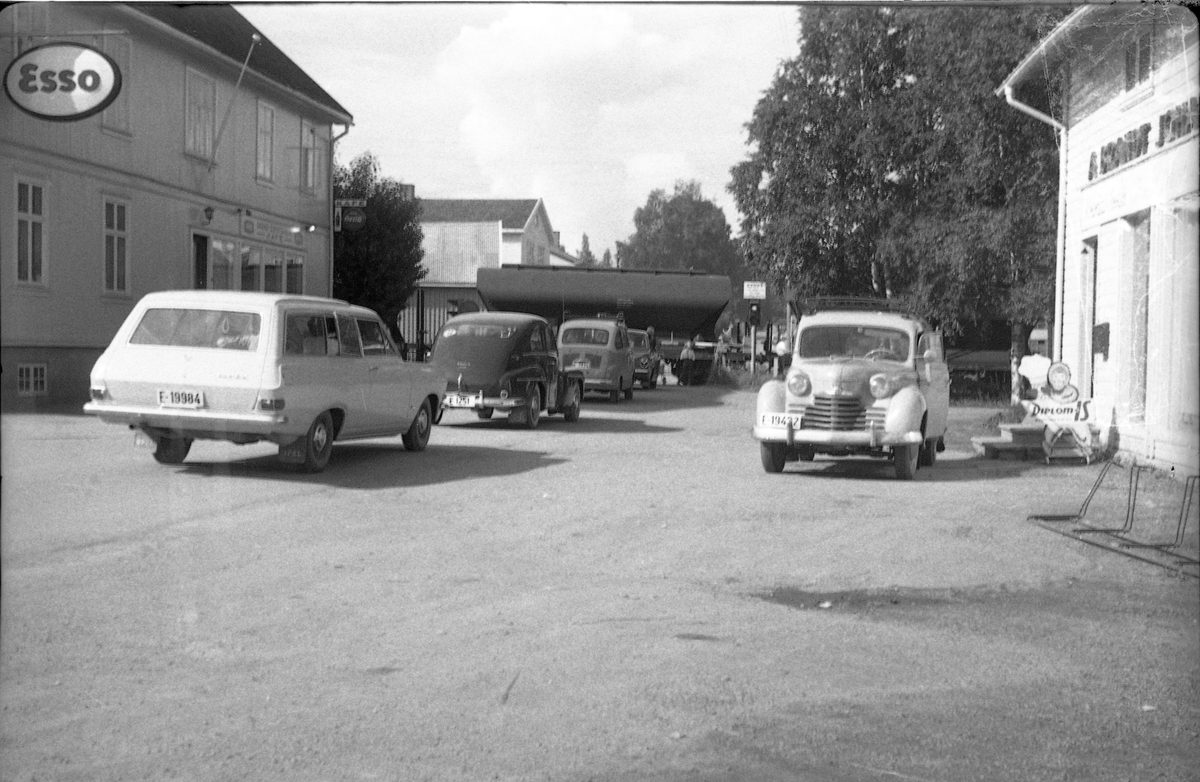 Gatebilde fra Lena, trolig 1962. Bildet er tatt inne fra Lena Foto & Radio i "Raubua", i retning mot jernbaneovergangen der et tog nettopp krysser. En rekke biler venter på at vegen skal åpnes igjen. De to nærmeste bilene er iflg. informant to generasjoner Opel: Til høyre (med front mot fotografen) en Opel Olympia varebil (kjennetegn: lakkert grill) årsmodell 1950-52, til venstre en Opel Caravan 1962-65.