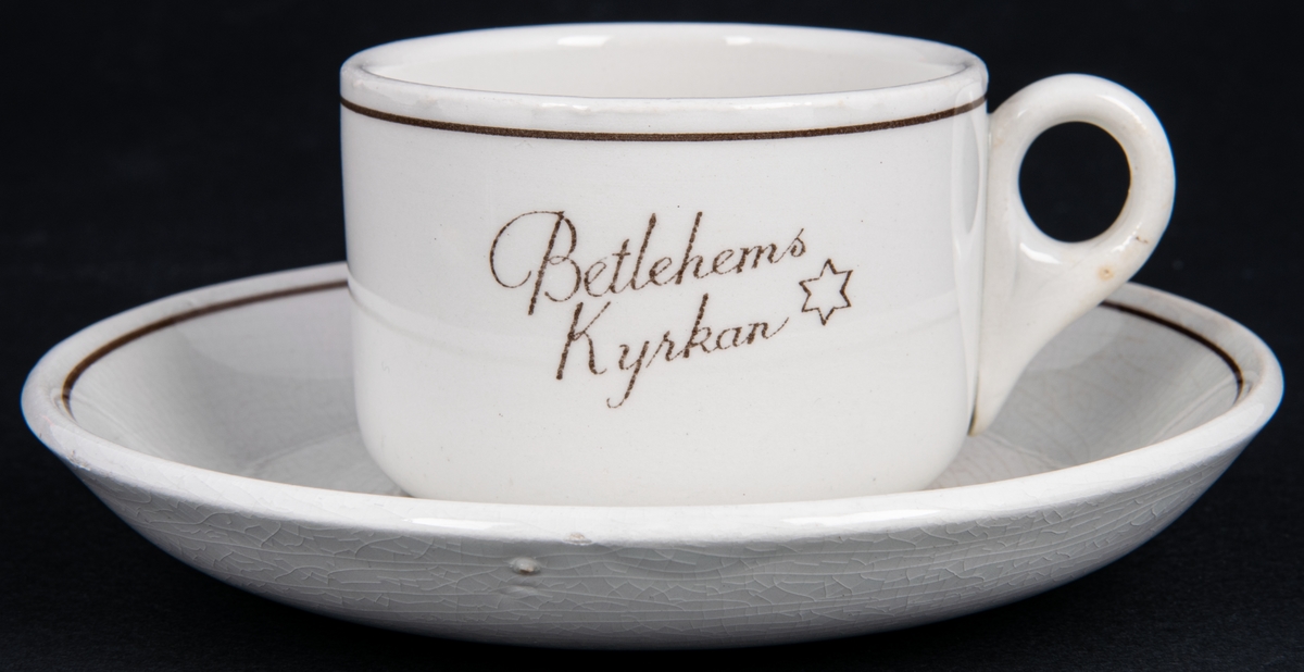 Kaffekopp med fat, vit glasyr med dekor i brunt med texten "Betlehems Kyrkan" samt en stjärna.