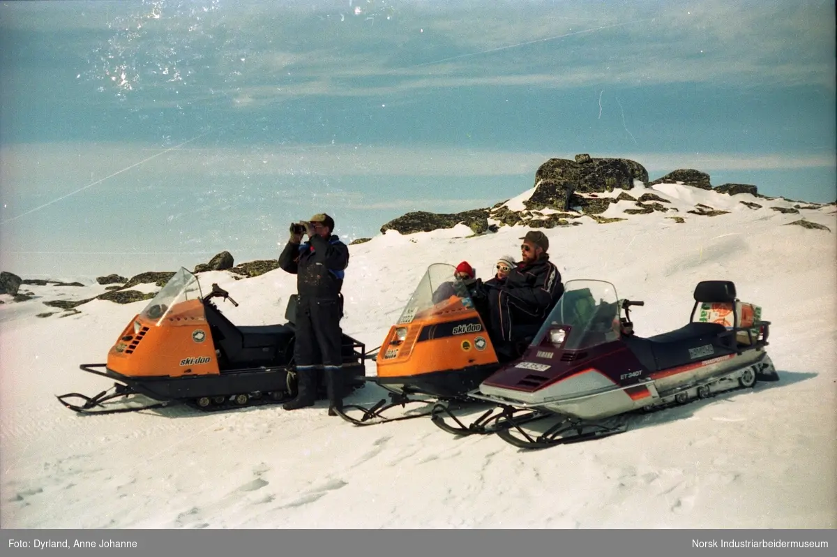 Snøskutetur i fjellet. Bjørn Dyrland står ved siden av skuter med kikkert. Bjørn Hagen med to personer til sitter på en skuter ved siden av