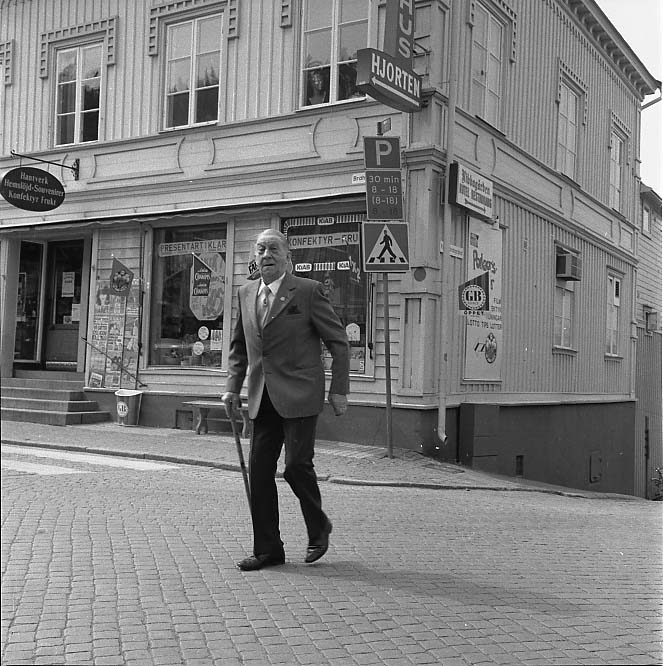 En äldre man, Dan Nicklasson, passerar över Brahegatan mot torget vid hörnet Brahegatan/Ribbagårdsgränd. Han har en käpp i handen och bär mörka byxor till ljusare kavaj med näsduk i bröstfickan samt slips. Bakom honom syns butiken på Brahegatan 39 med reklam för presentartiklar, konfektyr, hantverk och hemslöjd med mera.