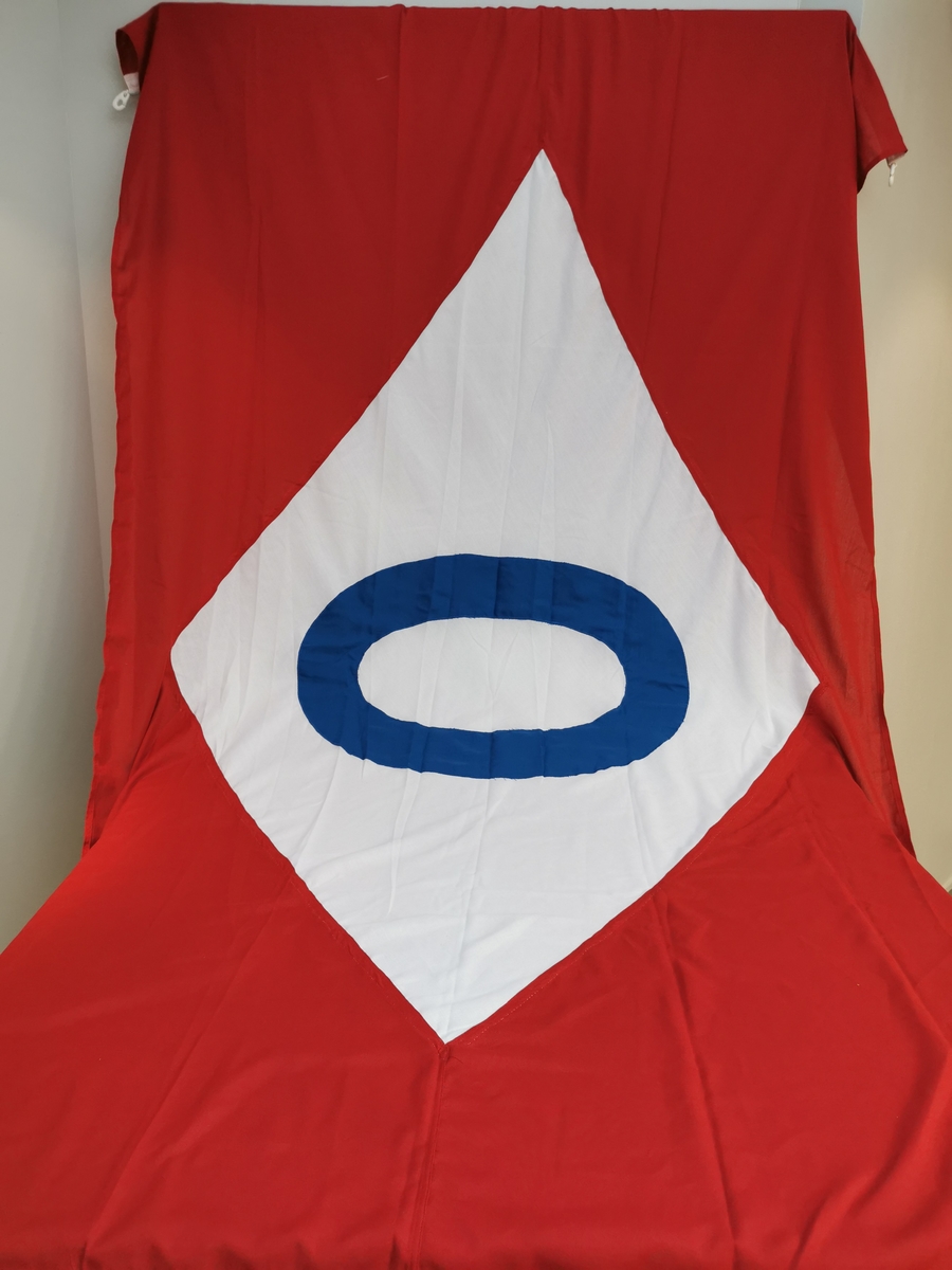 Rødt rektangulært flagg med en hvit skråstillt firkant midt på med en blå O. Flagget har antageligvis ikke blitt tatt i bruk. To flaggkroker i plast.