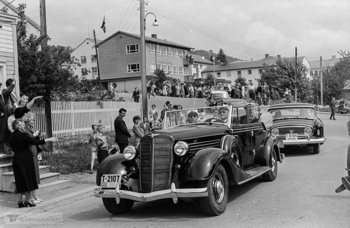 "Kongebesøket 24.06.1958".Kirkebakken i bakgrunnen..Lars Bugge sin konfeksjonsfabrikk i bakgrunnen med gammlegjemmet..T-2107 er Buick 1935, drosje T-8785 er Nash 1955.