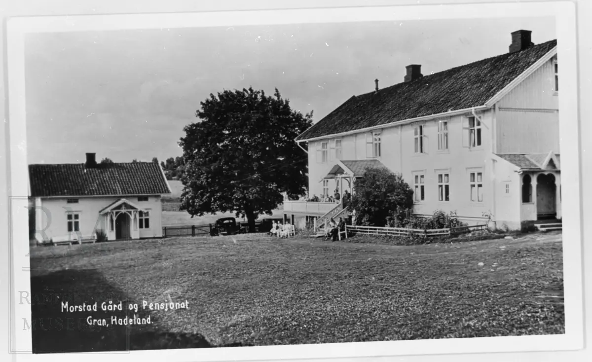 Tunet på Morstad vestre med en stor og en liten bygning. På verandaen og på bakken foran huset sitter det folk. En bil er parkert ved et stort tre.