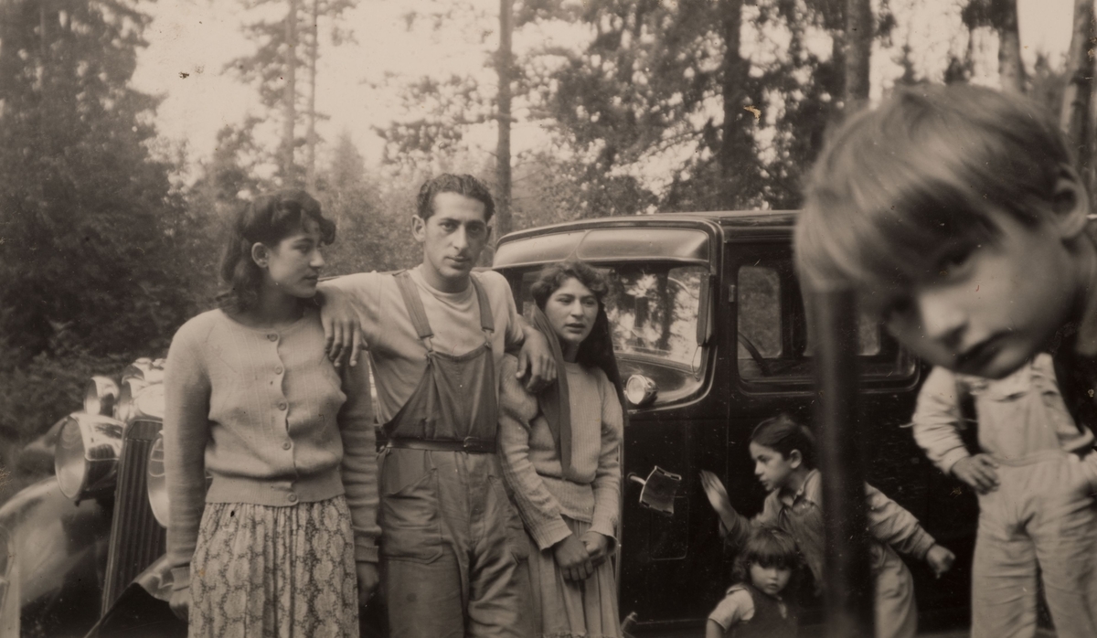 En familjen samlad för fotografering vid bilen. Ett barn kikar fram och skymmer en del av motivet. I bakgrunden syns barrskog. Bilden är tagen i juli 1950 i Storvik.