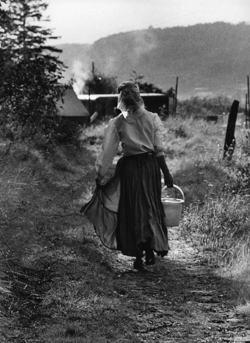 En romsk kvinna går på en stig bärandes en hink med vatten. Hon bär en dikhlo över håret och en lång kjol.