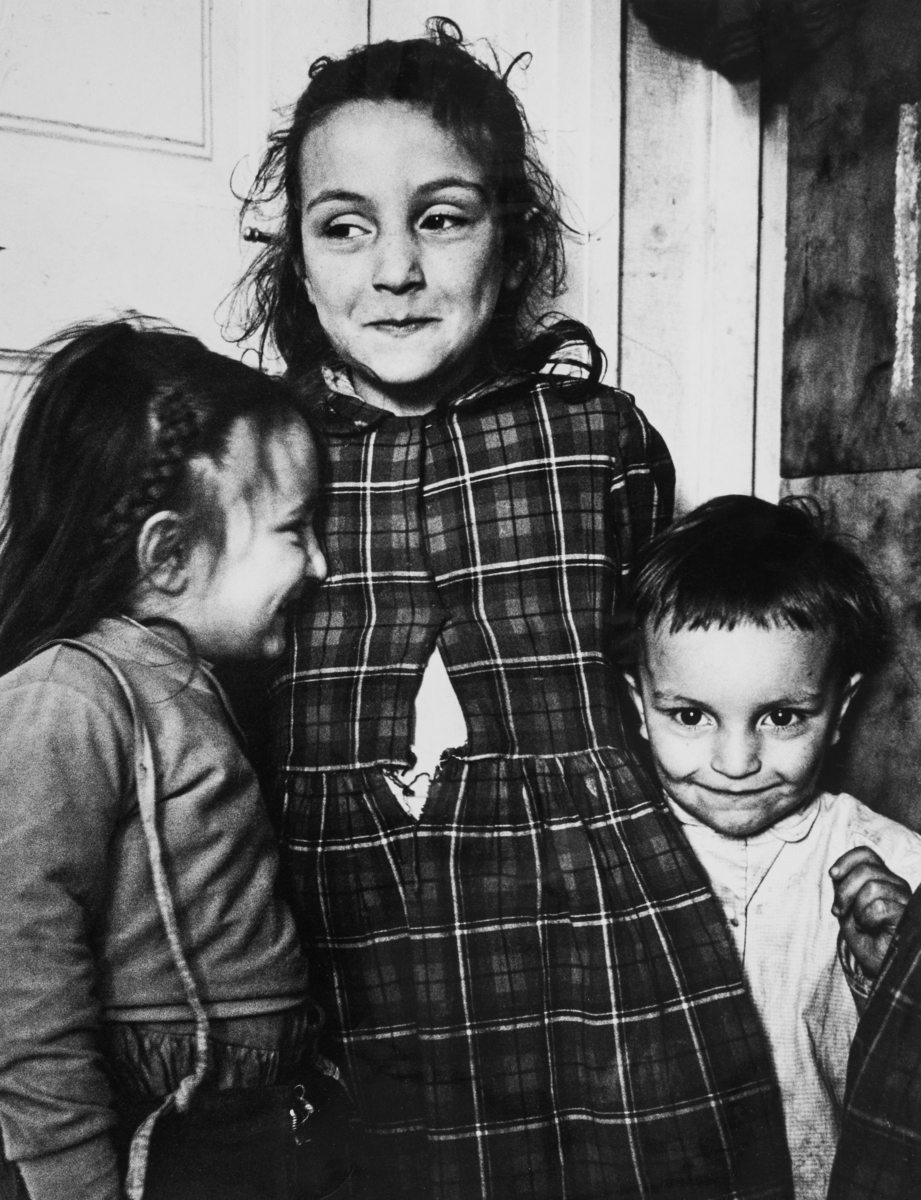 Tre romska barn står intill varandra vid en dörr. Det äldsta barnet bär en rutig klänning. Vid hennes högra sida kikar ett yngre barn fram och ett lite äldre barn står vid hennes vänstra sida med ansiktet mot hennes bröstkorg.