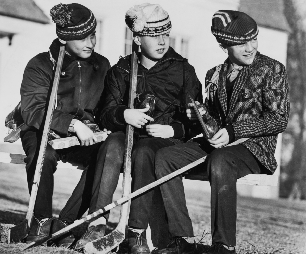 Tre pojkar sitter med skridskorna redo i famnen och mössorna på sned.