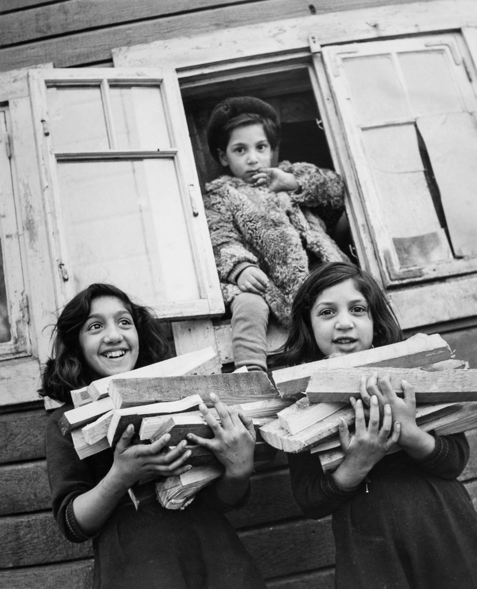 Två flickor ler mot kameran med famnarna fulla med ved. Bakom dem kikar ett yngre barn ut genom ett fönster. Bilden är tagen i Sköndal, som brukar beskrivas som det första av de uppsamlingsläger som Stockholms Stad kom att iordningsställa i mitten av 1900-talet.