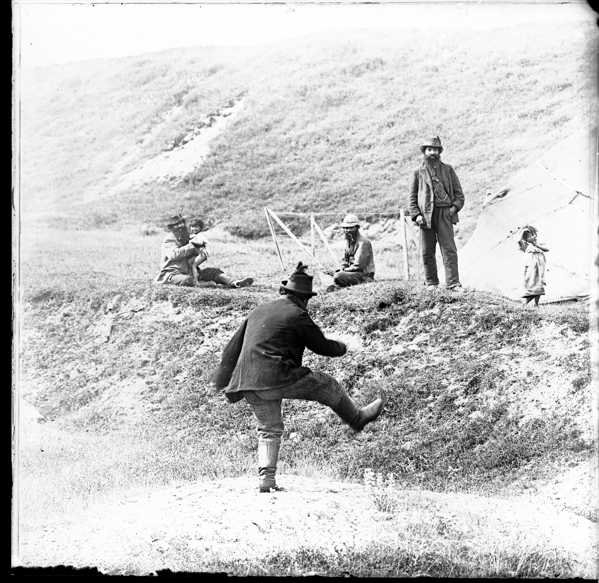I en backe har ett romskt sällskap slagit läger. En man hoppar och skojar. Några ytterligare män ser pån honom tillsammans med några barn. I bakgrunden syns ett tält.