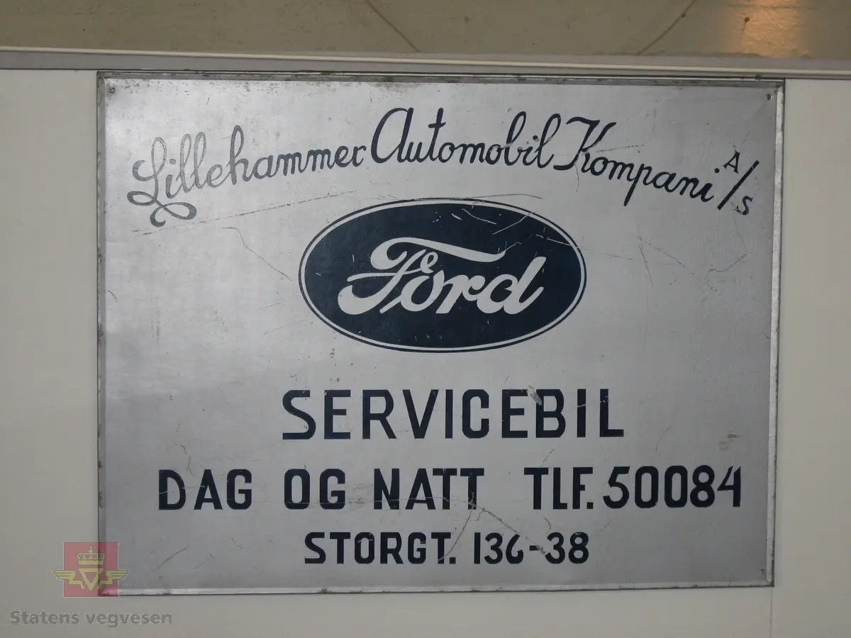 Rektangulært navneskilt i metall, med teksten " Lillehammer Automobil Kompani A/S SERVICEBIL DAG OG NATT TLF. 50084 STORGT. 136-38" påført med blå bokstaver på grå bakgrunn og Ford-emblemet midt på skiltet.