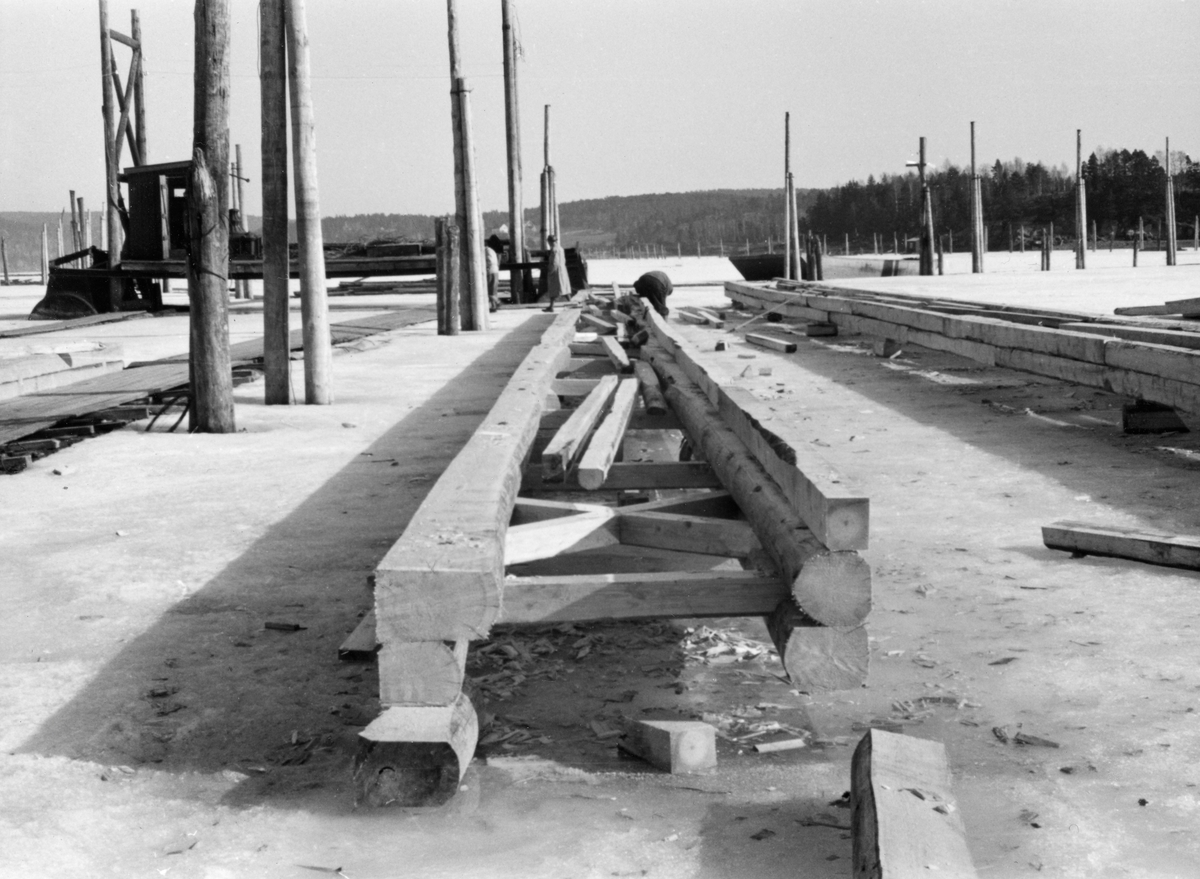 Bygging av nye ganglenser ved lenseanlegget på Eidet i Tune i Østfold vinteren 1953.  Arbeidet ble utført på isen på innsjøen Visterflo, ved sjølve lenseanlegget, der disse konstruksjonene skulle brukes.  Rammekonstruksjonen, som det ble arbeidet med her, besto av tømmer som var rettskåret på tre kanter og boltet sammen i anslagsvis 20-30 meters lengde.  Rammene var cirka en meter brede, med de nevnte sammenboltete tømmerkonstruksjonene på langsidene og med dels rette, dels krysslagte stag av tre mellom.  Konstruksjonen lå på kabber på isen, og ved den fremre enden av den påbegynte ganglensa som ligger sentralt i bildeflata ser vi en lensearbeider som sto bøyd over arbeidet sitt.  Til venstre for ham skimter vi en mann i frakk og hatt, antakelig en funksjonær.  Arbeidsstedet er omgitt av pælerekker som stikker opp gjennom isflata, og i mellomgrunnen til venstre i bildet skimter vi ei av de to mosemaskinene på Eidet.  I bakgrunnen ser vi lave, skogkledde åser. 